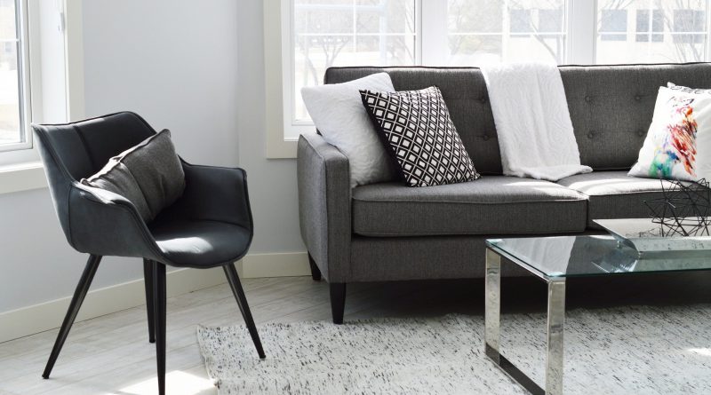 Skromne mieszkanie - krzesło, sofa i stolik. Biały dywan i białe ściany, szare meble. Za sofą znajduje się okno, widok na jakiś budynek